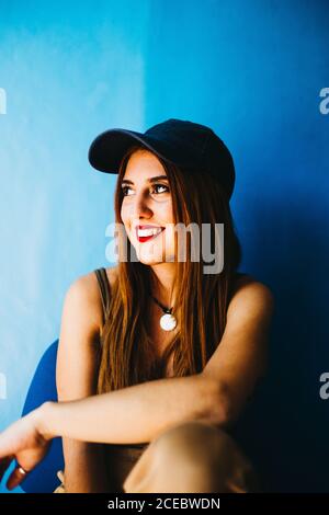 Giovane donna dai capelli rossi allegri in cappellino e canotta guardando lontano e seduto vicino al muro blu Foto Stock