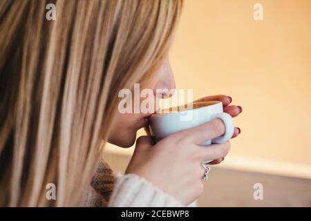 Vista laterale Crop attraente signora in maglione che tiene tazza di†caldo bere vicino alle labbra Foto Stock