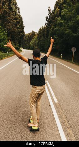 Vista posteriore di un giovane uomo a piedi nudi che mostra il segno shaka e. equitazione skateboard lungo la strada di campagna Foto Stock