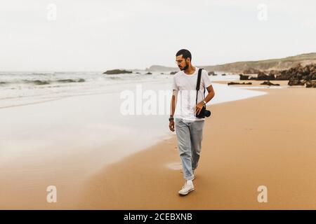 Attraente giovane uomo con macchina fotografica che cammina sulla spiaggia di sabbia e guardando l'acqua di mare mentre si viaggia nella natura Foto Stock