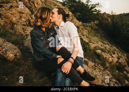 Giovane uomo che bacia la ragazza in guancia mentre si siede insieme pendio di montagna rocciosa Foto Stock