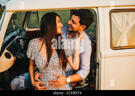 Giovane uomo felice e donna baciare e abbracciare mentre si trova in piedi a furgone vintage bianco in natura Foto Stock