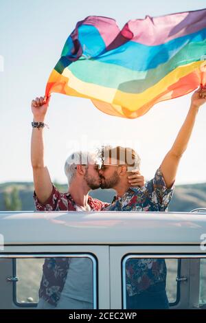 Due bei uomini che baciano e tengono la bandiera luminosa di LGBT mentre in piedi all'interno furgone con tetto aperto in campagna su soleggiato giorno Foto Stock