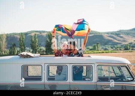 Due bei uomini che baciano e tengono la bandiera luminosa di LGBT mentre in piedi all'interno furgone con tetto aperto in campagna su soleggiato giorno Foto Stock