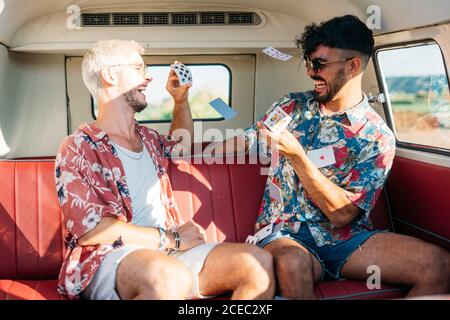 Due giovani allegri ragazzi ridendo e gettando carte da gioco mentre seduto sul sedile del passeggero del furgone retrò durante il viaggio in natura Foto Stock
