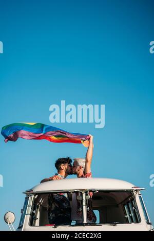 Due bei uomini che baciano e tengono la bandiera luminosa di LGBT mentre in piedi all'interno furgone con tetto aperto in campagna su soleggiato giorno Foto Stock