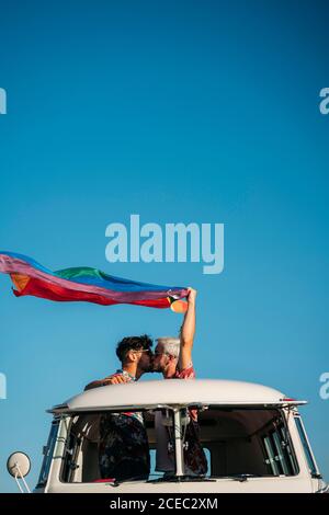 Due bei uomini che baciano e tengono la bandiera luminosa di LGBT mentre in piedi all'interno furgone con tetto aperto in campagna su soleggiato giorno Foto Stock