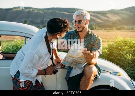 Due ragazzi con fotocamera in piedi vicino a auto retrò e guardando la mappa mentre si viaggia in natura insieme Foto Stock