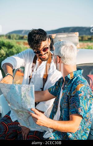 Due ragazzi con fotocamera in piedi vicino a auto retrò e guardando la mappa mentre si viaggia in natura insieme Foto Stock