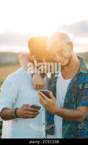 Due uomini sorridenti che abbracciano e navigano gli smartphone mentre si trovano in piedi bella campagna insieme Foto Stock