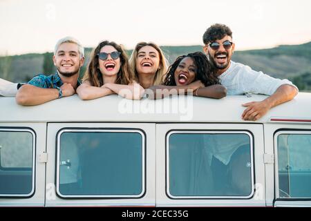 Gruppo di diversi giovani amici in piedi in berlina sul tetto di pulmino d'epoca e guardando la macchina fotografica mentre si passa il tempo in natura insieme Foto Stock