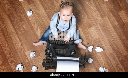 Dall'alto adorabile ragazzino seduto a macchina da scrivere con fogli di carta sul pavimento. Foto Stock