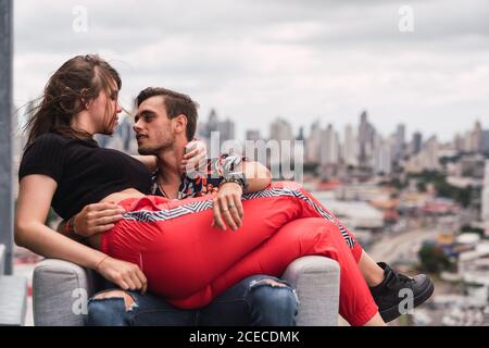 Attraente giovane uomo che abbraccia bella donna mentre si siede in comoda poltrona su sfondo sfocato della città moderna nel giorno nuvoloso Foto Stock
