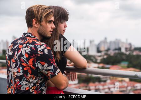 Vista laterale di un giovane ragazzo in un elegante vestito che abbraccia una bella donna dal retro mentre si alza sul balcone e si ammira la vista della città moderna insieme Foto Stock