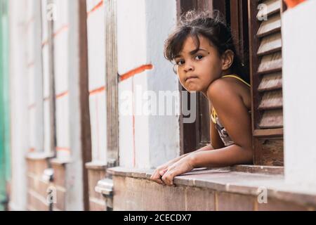 LA HABANA, CUBA - NOVEMBRE, 6 , 2018: Ragazzina pensante appesa fuori dalla finestra dell'edificio con facciata bianca a Cuba Foto Stock