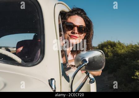 Felice giovane donna in occhiali da sole alla moda guardando fuori dalla finestra di auto godendo la luce del sole estivo contro gli alberi verdi e il cielo blu Foto Stock