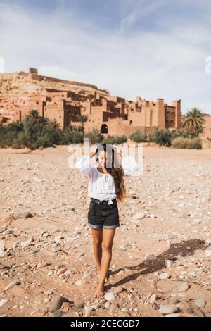 Giovane donna riccia orientale aspetto camminare sullo sfondo di kasbah Ait-ben-Haddou. Viaggio in Marocco, Ouarzazate. Vacanze estive, concetto di stile di vita di viaggio. Foto Stock