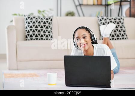 Ritratto della giovane donna nera positiva che indossa le cuffie quando si sdraia sul pavimento e lavorando su un computer portatile a casa Foto Stock