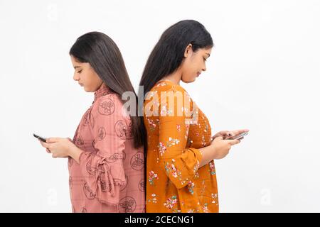 Due giovani ragazze indiane che si sono occupate dei loro smartphone, in piedi contro lo sfondo bianco dello studio. Foto Stock