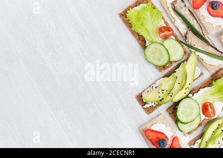 Set di panini di stagione biologica su croccanti a grani marroni secchi con verdure fresche, frutta matura, conserve di pesce, formaggio cremoso su tavolo di legno bianco Foto Stock