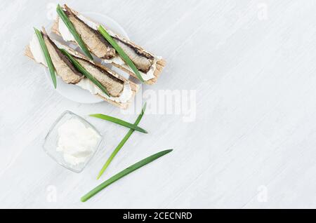 Sana colazione dietetica di pane di grano integrale con acciughe, formaggio cremoso, cipolla verde germogliata su sfondo di legno bianco, vista dall'alto, copia Foto Stock