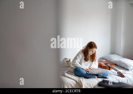 Bella giovane donna sorridente e navigante moderno laptop mentre si siede su comodo letto vicino alla chitarra acustica Foto Stock
