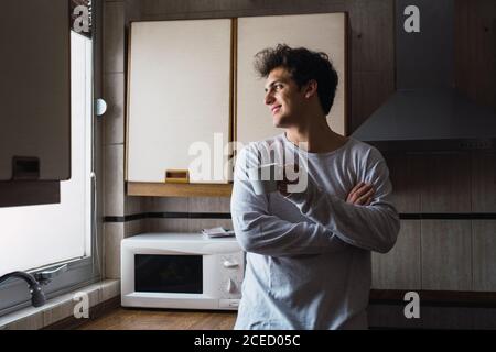 Giovane allegro in piedi con una tazza di caffè e guardando la finestra in cucina. Foto Stock