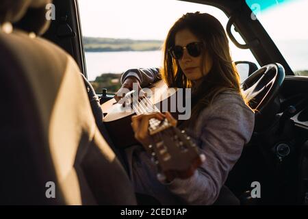 Attraente giovane donna che suona la chitarra e seduto in auto in natura. Foto Stock