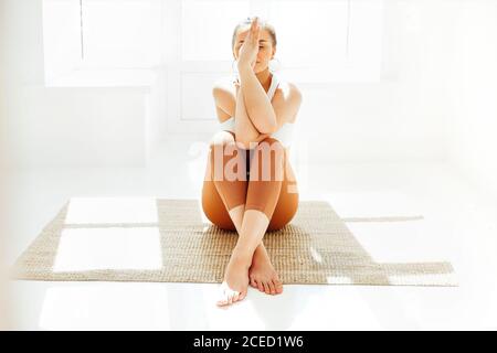 Giovane donna in abbigliamento sportivo seduta con gambe incrociate e mani di mudra sul tappetino e facendo yoga mentre si pratica la consapevolezza Foto Stock