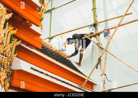 Da sotto l'uomo decoratore appeso sul cantiere e tetto di pittura di edificio asiatico. Foto Stock