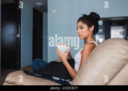 Bella brunetta Ispanica Donna tenere forchetta con fetta di frutta e sedersi sul divano con ciotola di kiwi e anguria Foto Stock