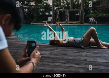 Fotografo irriconoscibile che scatta foto di Asian Woman sdraiata al bordo della piscina. Foto Stock