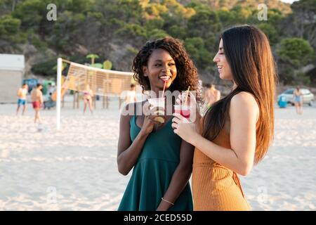 Diverse giovani amiche che si rilassano sulla spiaggia con bevande in tazza e ridendo mentre chiacchiera alla luce del tramonto Foto Stock