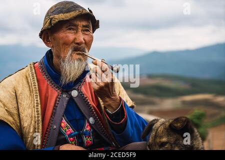 DONGCHUAN RED, YUNNAN, CINA - 14 GIUGNO 2018: Uomo anziano rilassato in abiti tradizionali luminosi seduti a terra fumando pipa di tabacco in legno e accarezzando il cane seduto?nelle vicinanze guardando via sullo sfondo offuscato delle terre rosse di Dongchuan nella provincia cinese di Yunnan Foto Stock