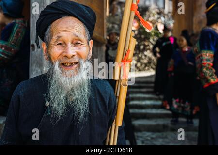 Allegro maschio invecchiato con barba grigia lunga in abiti scuri In piedi con strumento musicale al muro su un vicolo cinese stretto E guardando la macchina fotografica con altre persone di etnia Miao Minoranza su sfondo offuscato nella provincia di Guizhou Foto Stock