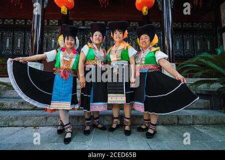 GUINZHOU, CINA - 14 GIUGNO 2018: Gruppo di belle donne cinesi della minoranza Miao che si trova in un edificio tradizionale in abiti da festival luminosi con ornamenti che tengono gonne e che guarda sorridente nella provincia di Guizhou Foto Stock