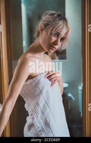 Affascinante giovane donna avvolto in bianco asciugamano strofinando faccia e guardando la macchina fotografica mentre in piedi nella porta del bagno dopo la doccia Foto Stock