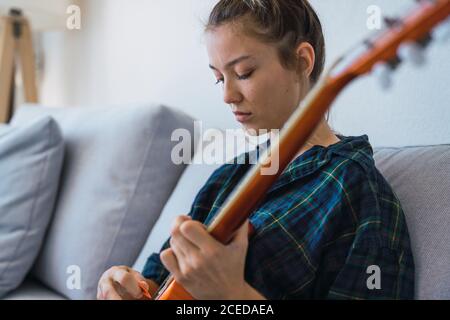 Attraente giovane donna seduta su comodo divano e utilizzando Pick per suonare la chitarra mentre si passa il tempo a casa Foto Stock