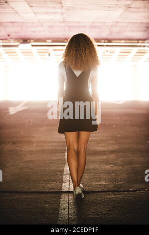 Vista posteriore di Slim Woman con ricci indossando abito nero con sneakers camminare nel parcheggio sotterraneo alla luce del sole Foto Stock
