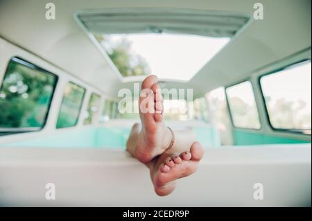 Gambe di irriconoscibile donna a piedi nudi sdraiata sul retro del sedile all'interno di un furgone d'epoca durante il viaggio nella natura Foto Stock