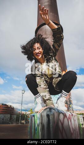 Da sotto di felice sportiva moderna Donna con ricci equilibrando su grungy recinto sorridendo alla macchina fotografica alla luce del sole Foto Stock