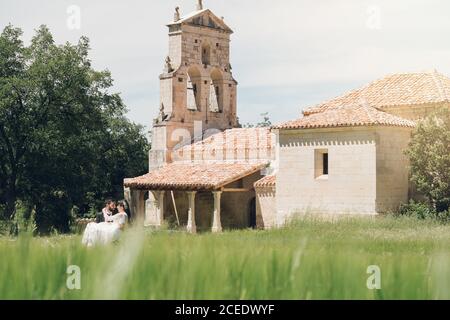 Giovane uomo in costume nero che tiene giovane donna in abito da sposa in braccia sullo sfondo di alberi verdi e vecchia chiesa di pietra al giorno di sole Foto Stock