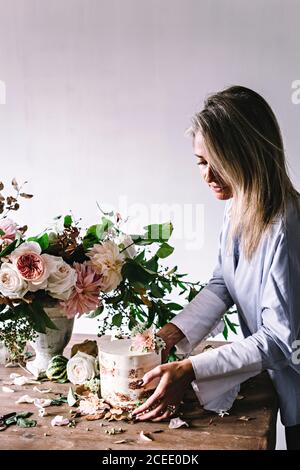 Vista laterale della signora posizionando il piatto con una gustosa torta decorata bloom bud sul tavolo di legno con mazzo di crisantemi, rose e rametti di piante in vaso tra Foto Stock