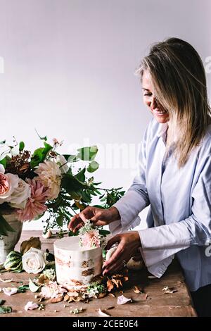 Vista laterale della signora posizionando il piatto con una gustosa torta decorata bloom bud sul tavolo di legno con mazzo di crisantemi, rose e rametti di piante in vaso tra Foto Stock