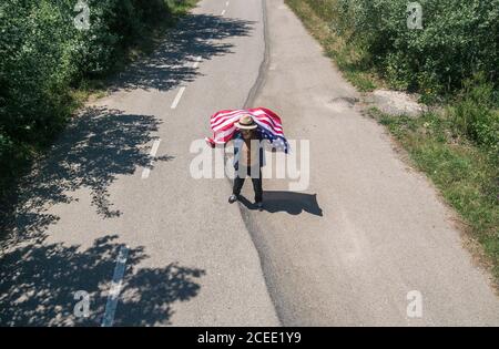 Uomo in cappello che cammina con la bandiera americana su una strada solitaria. Giornata speciale per festeggiare il 4 luglio Foto Stock