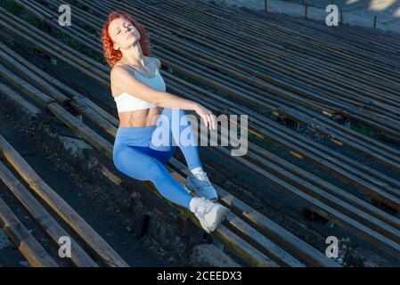 giovane sportivo dai capelli rossi si siede su una panchina di legno di uno stadio sportivo ai raggi del sole che tramonta su uno sfondo scuro. Sport, stile di vita attivo Foto Stock