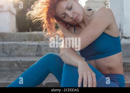 Ritratto di una giovane bella atleta dai capelli rossi che ascolta la musica con le cuffie, seduto sui gradini, sullo sfondo dell'ambientazione Foto Stock