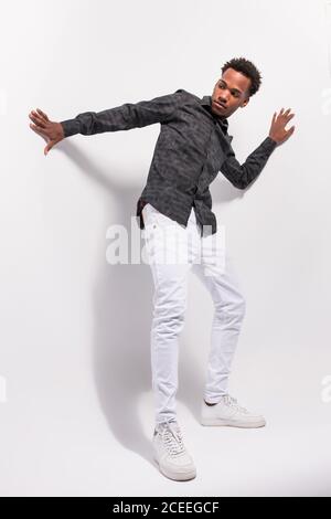 Da sotto colpo di trendy afroamericano giovane in bianco denim e camicia su sfondo bianco in postura Foto Stock