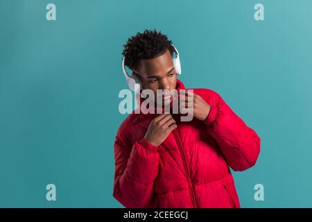 Elegante uomo afro-americano con cuffie bianche e cappotto rosso invernale su sfondo blu Foto Stock