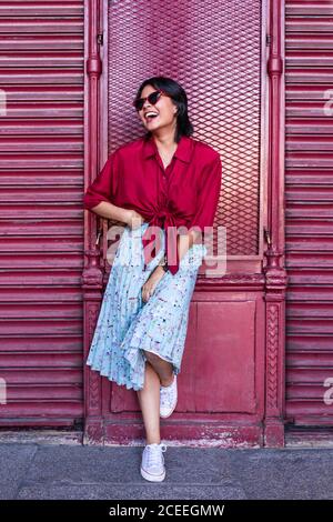 Bella giovane donna in abito alla moda ridendo e guardando via mentre si appoggia sul muro di borgogna sulla strada della città Foto Stock